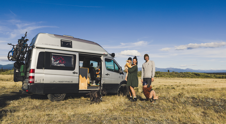 The roadtyping family stands together with their dog in front of their camper, surrounded by nature